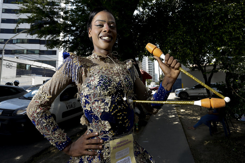 A foto mostra Priscilla Tawanny, mulher negra e jovem em um ambiente externo durante o dia, segurando duas balizas de banda marcial. A pessoa está vestida com uma fantasia elaborada, adornada com lantejoulas e miçangas que criam padrões intrincados e brilham à luz. A fantasia inclui mangas longas e cobre totalmente o torso, sugerindo que pode fazer parte de uma roupa de carnaval ou festiva. Ao fundo, há carros estacionados e árvores,