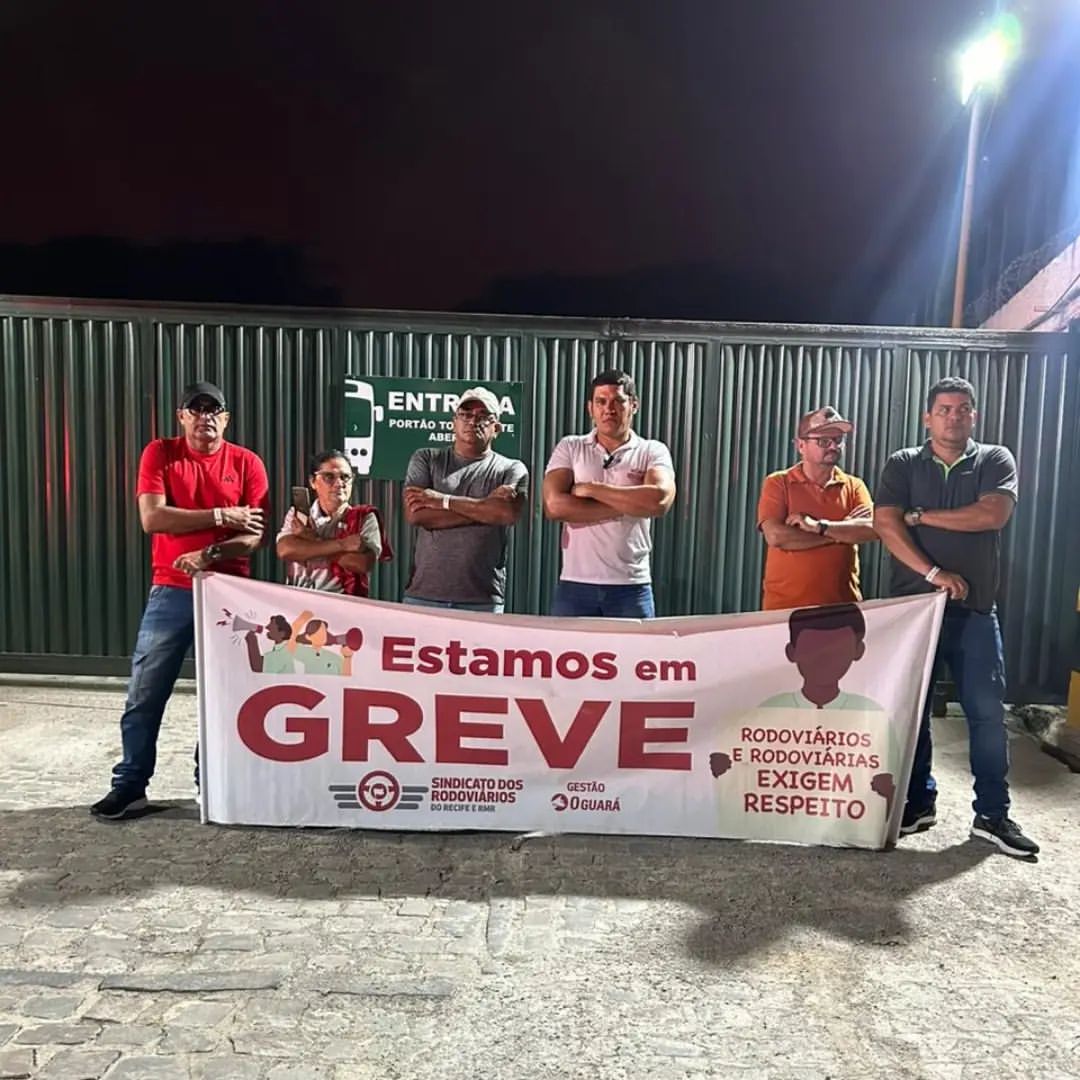 A imagem mostra um grupo de cinco pessoas em pé atrás de uma faixa. A foto foi feita à noite. A faixa tem um texto importante que indica o propósito da reunião. As pessoas parecem estar participando de uma manifestação ou protesto, como sugerido pela faixa que diz “Estamos em GREVE”. Esta frase é central para entender o contexto da imagem, indicando que essas pessoas provavelmente são trabalhadores participando de uma greve trabalhista.