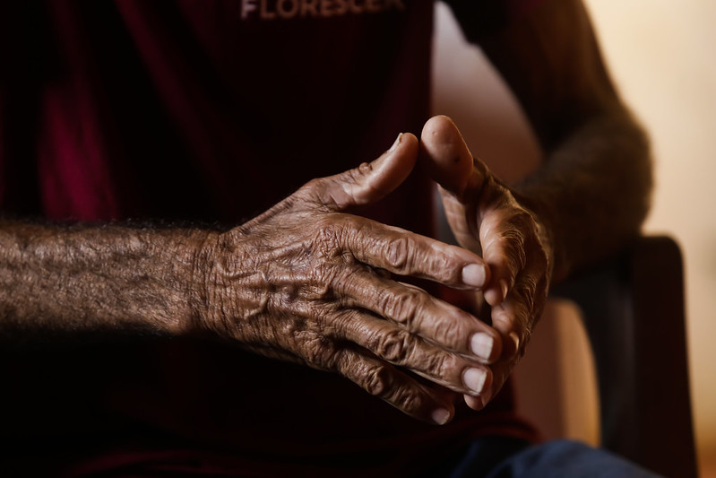 Foto de mãos de um homem idoso, bastante enrugadas com a pele curtida e bronzeada pelo sol. As pontas dos dedos das duas mãos estão se tocando. O homem usa uma camisa vermelho escuro, única coisa que se vê na fotografia, feita em um ambiente escuro.