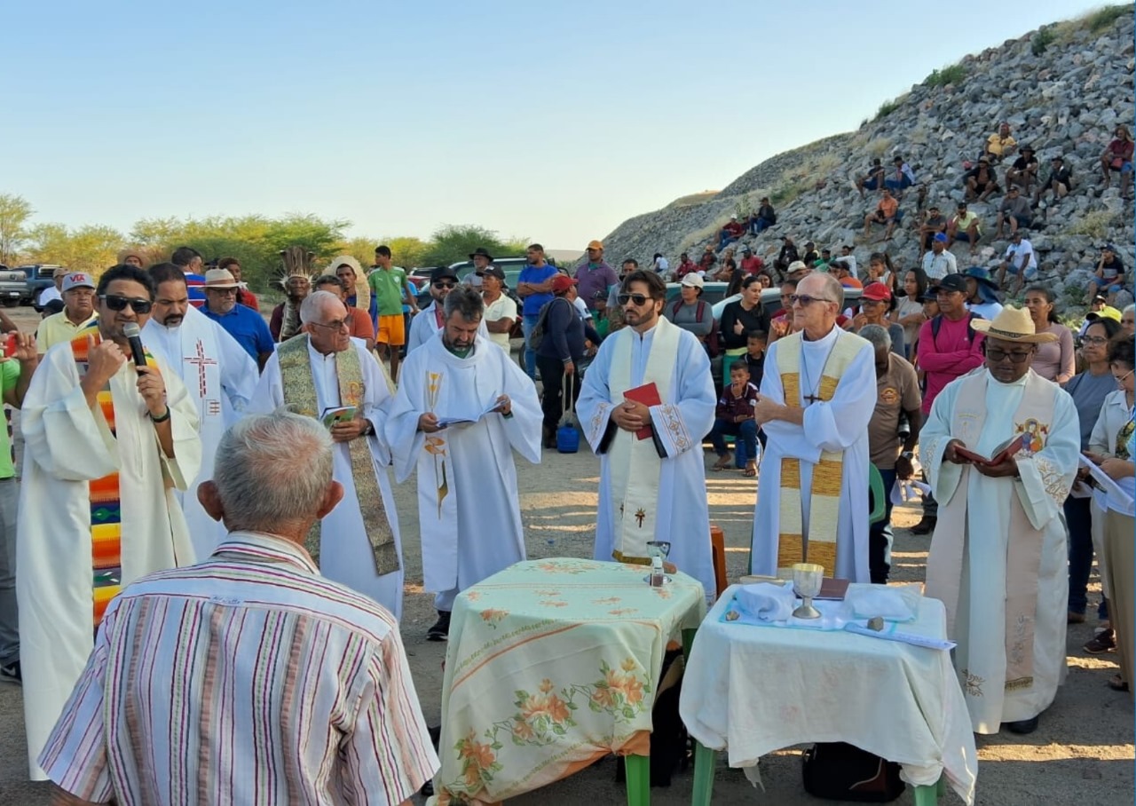 Grupo de sete sacerdotes católicos, todos homens, vestidos com batinas e estolas (trajes ritualísticos) reunidos ao ar livre em torno de duas pequenas mesas cobertas com toalhas claras. Eles estão celebrando uma missa campal, tendo em torno de si uma pequena multidão que acompanha a cerimônia.