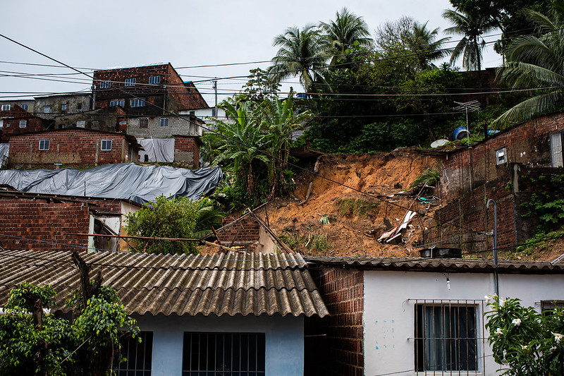 A imagem mostra uma área urbana em um terreno íngreme. Há várias casas construídas próximas umas das outras, com estilos arquitetônicos variados e diferentes estados de conservação. No primeiro plano, há uma casa com telhado azul e paredes brancas. Atrás dessa casa, há uma encosta íngreme e sem vegetação, com sinais visíveis de erosão do solo. Acima da encosta, mais casas podem ser vistas, densamente agrupadas, feitas de materiais como tijolo, concreto e chapas de metal ondulado. Alguns telhados estão cobertos com lonas azuis. O céu está nublado, sugerindo que pode ser um dia nublado.