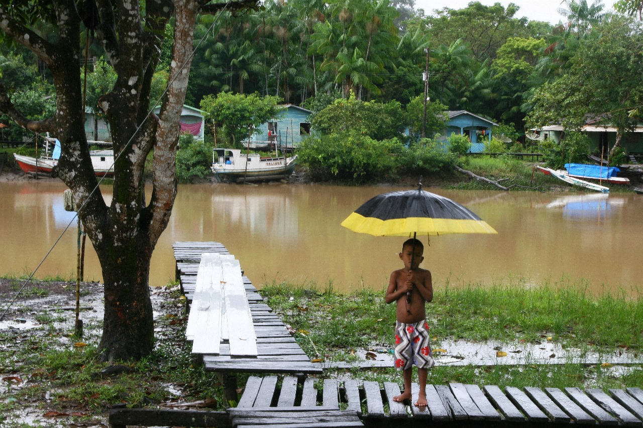 A foto mostra uma criança de costas, em pé em um cais de madeira, segurando um guarda-chuva amarelo e preto. A criança está olhando para um rio de águas barrentas. Na margem oposta do rio, há várias casas coloridas sobre palafitas e barcos atracados ao longo da margem. O céu está nublado, sugerindo que pode estar chovendo ou que choveu recentemente. A cena parece retratar a vida cotidiana em uma comunidade ribeirinha