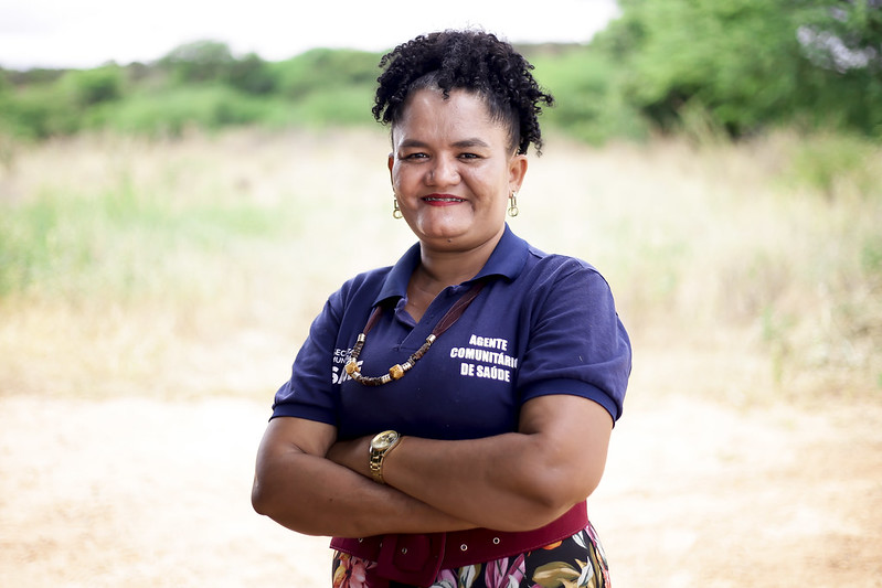 Foto de Viviane Paiva, fotografada acima da cintura em um ambiente ao ar livre. Ela é uma mulher negra, madura, de cabelos curtos encaracolados. Ela está usando uma camisa polo azul escura onde se lê agente comunitário de saúde, tendo um colar artesanal marrom de fibras grossas. Ela também está de brincos de metal, relógio no pulso esquerdo, com os braços cruzados no abdômen. É possível ver que ela usa uma saia ou calças estampadas.