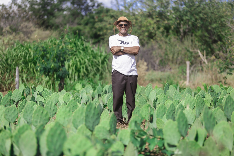 A imagem mostra um homem negro de meia idade de pé no meio de um campo de cactos verdes com folhas largas. O homem usa um chapéu de couro e óculos escuros. Ele está vestindo uma camiseta branca com um gráfico preto, calças escuras e sapatos. Seus braços estão cruzados sobre o peito e ele tem um sorriso largo, bastante aberto. Ao fundo, há árvores e arbustos sob um céu claro.