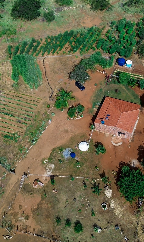 A imagem mostra uma vista aérea de uma paisagem rural. No centro, há uma casa com telhado vermelho, cercado por várias áreas de vegetação, incluindo fileiras de plantações e árvores espalhadas. Ao lado do edifício, há duas estruturas circulares, possivelmente tanques de água, uma azul e outra branca, com tubulações ligando essas estruturas ao imóvel. O terreno é predominantemente verde e marrom, indicando áreas cultivadas e caminhos.