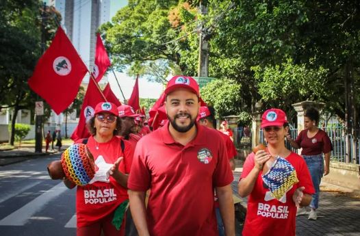 A imagem mostra um grupo de pessoas participando de uma manifestação ou evento público. Caminhando à frente de duas mulheres jovens, está Tomás Agra, um homem jovem, branco, de barba curta e bem feita, usando camisa polo vermelha e um boné vermelho com o símbolo do MST. As pessoas estão segurando bandeiras vermelhas com um círculo branco e um símbolo preto no meio, que é característico do Movimento dos Trabalhadores Rurais Sem Terra (MST) no Brasil. Algumas pessoas estão vestindo camisetas vermelhas com o texto “BRASIL POPULAR” em letras brancas e grandes, sugerindo que este é um evento relacionado a questões sociais ou políticas brasileiras. Além disso, uma pessoa está segurando um instrumento musical semelhante a um chocalho decorado com contas coloridas. Ao fundo, há árvores e prédios altos e modernos, indicando que o evento está ocorrendo em um ambiente urbano.