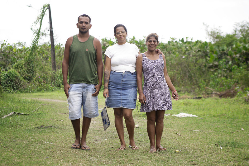 Foto de Carlos César da Silva, Jéssica Ferreira e Elania Maria da Silva em pé diante de uma área de matagal. Ele é um homem jovem negro, de cabelos cursos, barba e bigode ralos, usando camiseta regata verde musgo, bermuda jeans e sandálias do tipo havaianas. Jéssica é uma mulher parda jovem, de cabelos presos por trás da cabeça, camiseta branca, saia jeans, e sandálias do tipo havaianas. Elania é uma mulher negra, de cabelos crespos castanhos, usando vestudo estampado nos tins cinza e branco, com sandálias do tipo havaianas.