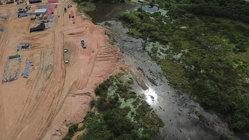 Foto aérea de aterro da Muribeca onde se vê o solo alaranjado da área aterradas avançando sobre o leito de um rio, com vegetação baixa à direita do curso d'água.