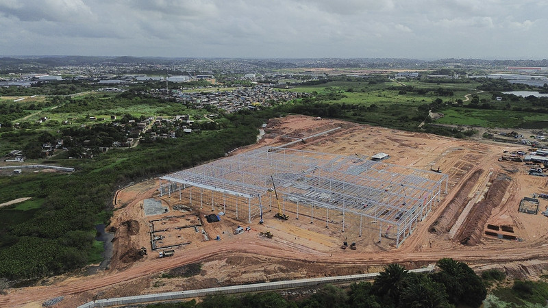 Foto aérea de um aterro na Muribeca, onde vê um extenso terreno plano de solo alaranjado no centro e à direita da imagem, com estrutura de metal sendo erguida no centro da foto. Há algumas construções ao fundo, e vegetação à direita e à esquerda da foto. O céu está nublado, com muitas nuvens cinzas.