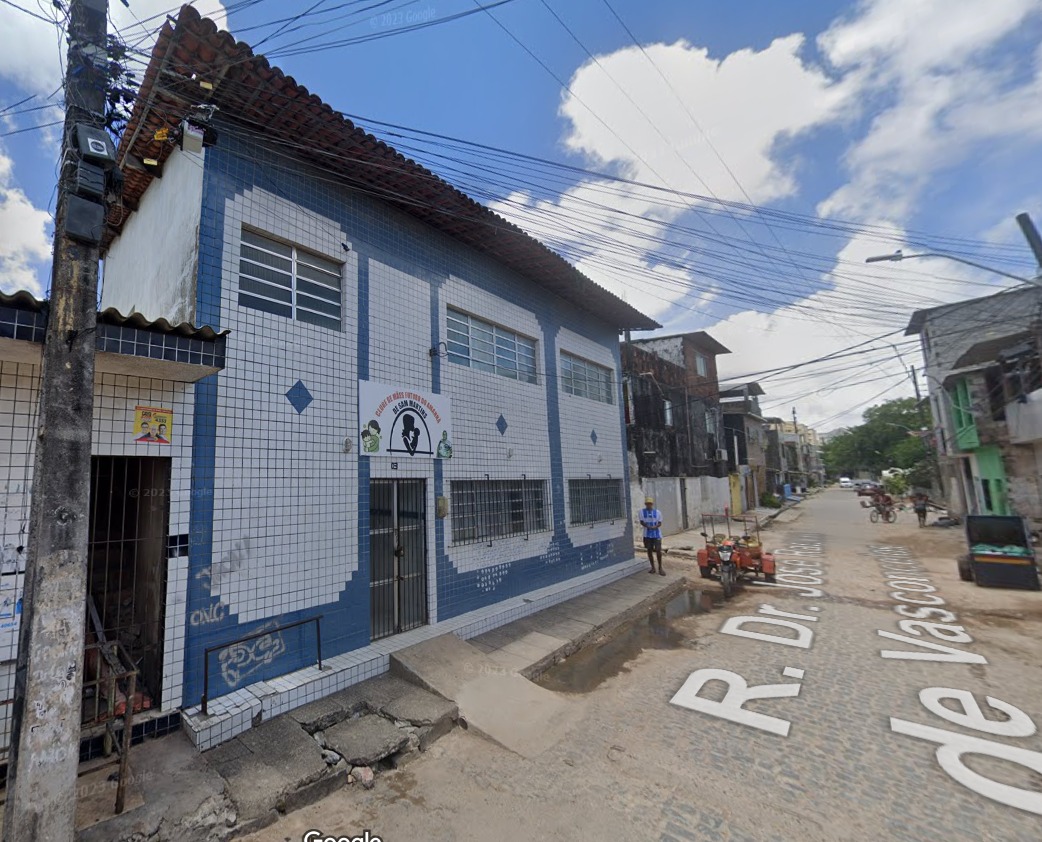 A imagem mostra um prédio de dois andares em uma esquina. O prédio tem uma fachada azul e branca com padrões de azulejos. Há vários fios elétricos passando acima da rua, conectados a um poste à esquerda do prédio. No andar térreo, há uma entrada com um design em arco acima dela, apresentando o que parece ser um logotipo ou emblema que inclui um globo e duas figuras humanas. No segundo andar, há janelas com grades de metal para segurança. À direita do prédio, há outra rua onde mais edifícios podem ser vistos em vários estados de conservação e com diferentes esquemas de cores.