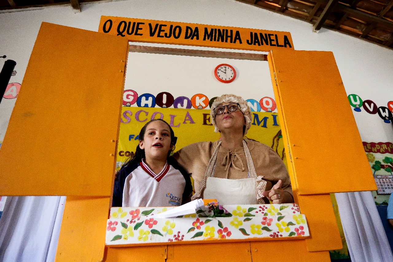 A imagem mostra um ambiente interno com uma grande moldura de janela montada na parede. No topo da moldura, há um texto em português que diz “O que vejo da minha janela”. Dentro dessa moldura, há duas pessoas - à esquerda, uma menina vestindo uma camisa de uniforme escolar, e à direita, uma mulher adulta usando óculos, avental estampado e uma peruca branca para aparentar ser mais velha. Ambas estão segurando folhas de papel, como se estivessem lendo um texto. A parede ao redor da moldura está decorada com trabalhos artísticos de crianças e recortes coloridos de papel, incluindo letras do alfabeto. Abaixo da moldura, há uma prateleira com vários pequenos objetos e mais materiais de arte espalhados. O teto do ambiente tem vigas de madeira, e há um relógio na parede que marca aproximadamente dez minutos após as dez horas.