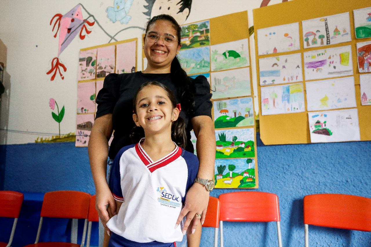 O ambiente na imagem é uma sala de aula. As paredes estão decoradas com desenhos e obras de arte coloridas, incluindo representações de animais, paisagens e vários designs abstratos. A parte inferior das paredes é azul, enquanto a parte superior é branca, com vários desenhos infantis fixados. Há cadeiras vermelhas visíveis no primeiro plano, sugerindo uma área de assentos para alunos ou participantes. Na sala de aula estão duas pessoas. Uma mulher adulta jovem, parda, de cabelos pretos compridos, usando óculos, blusa preta e relógio dourado no pulso esquerdo. À sua frente, encostada em seu corpo está uma menina branca, de cabelos pretos lisos, usando blusa de uniforme escolar branca com mangas azuis e gola vermelha.