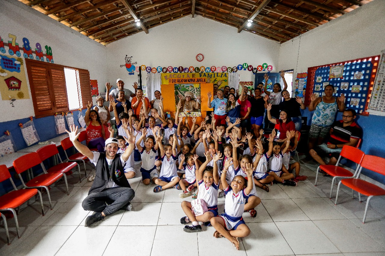 A imagem mostra uma sala de aula com várias crianças e alguns adultos. As crianças estão usando uniformes combinando, com camisas brancas e calças ou saias azul-marinho. Elas estão sentadas em cadeiras vermelhas dispostas em fileiras, com as mãos levantadas, como se estivessem respondendo a uma pergunta ou se voluntariando para uma atividade. No primeiro plano, uma criança está de pé com os braços levantados. Um adulto está ajoelhado no chão em frente a essa criança, fazendo um sinal de paz com a mão. Outros adultos estão ao fundo da sala, alguns também com as mãos levantadas. As paredes estão decoradas com pôsteres educativos coloridos e trabalhos artísticos.