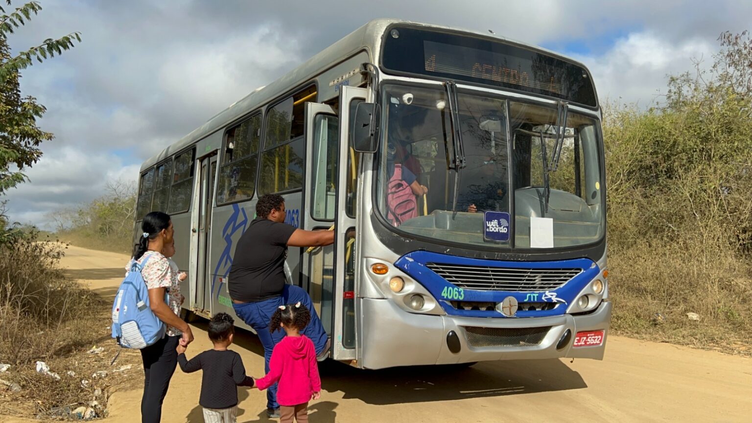 A imagem mostra um ônibus em uma estrada de terra, com vegetação dos dois lados. O ônibus é prateado e azul, com a porta da frente aberta e uma pessoa de camisa rosa visível através do para-brisa. Há três pessoas em pé ao lado da porta: um adulto ajudando outro adulto a subir no ônibus, e uma criança segurando a mão do segundo adulto. O céu está parcialmente nublado, sugerindo um ambiente rural ao ar livre.