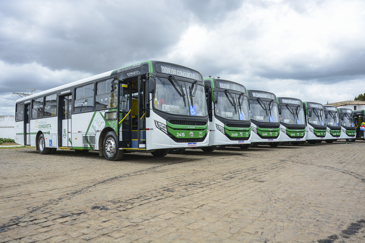 A imagem mostra uma frota de ônibus estacionados em um pátio ou área de estacionamento. Há seis ônibus visíveis, alinhados diagonalmente da parte inferior esquerda para a parte superior direita da imagem. Os ônibus têm um esquema de cores verde e branco, com detalhes azuis perto da área do motorista. Cada ônibus possui um letreiro de destino acima do para-brisa, embora o texto não seja legível. O céu está nublado, sugerindo que pode ser de manhã cedo ou fim de tarde.