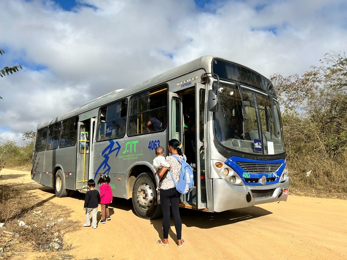 A imagem mostra um ônibus prateado em uma estrada de terra, cercado por vegetação seca, sob um céu azul claro. O ônibus tem gráficos azuis e cinza, e o número “4093” é visível na frente e na lateral. Três pessoas estão em primeiro plano: um adulto vestindo roupas escuras e carregando uma mochila, caminhando em direção à porta aberta do ônibus, e duas crianças, uma vestida de vermelho e outra de azul, também indo em direção ao ônibus. O ambiente sugere uma área rural ou menos urbanizada