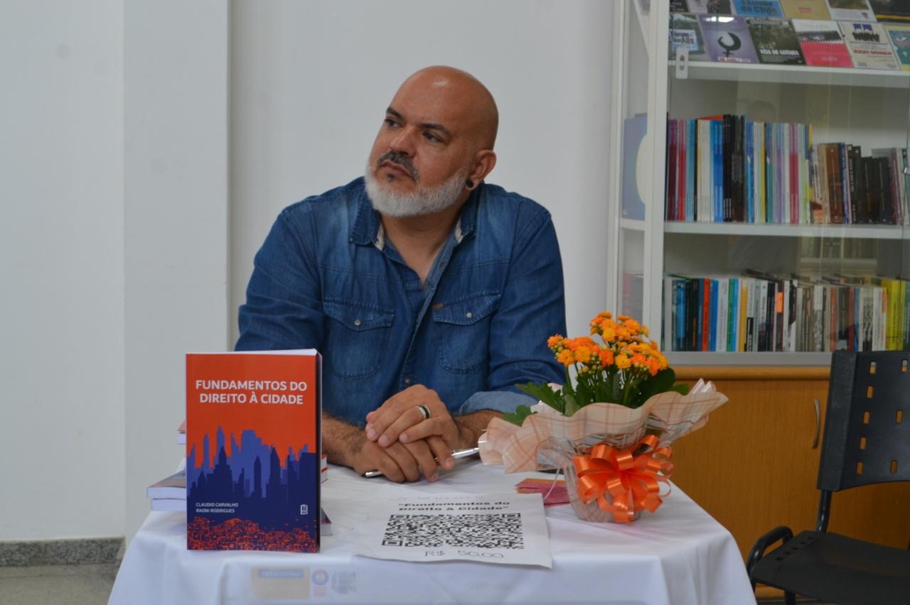 Foto de Cláudio Carvalho, homem branco de meia idade, de cabeça raspada, barba branca e bigode grisalho, usando camisa jeans de mangas compridas. Ele está sentado à uma pequena mesa coberta por toalha branca onde um exemplar do livro Fundamentos do Direito à Cidade, de capa laranja e azul, está colocado à frente da mesa. Ao lado o livro, há uma pequena cesta de flores laranjas. A foto foi feita em ambiente fechado, com uma pequena estante de livro à direita da imagem.