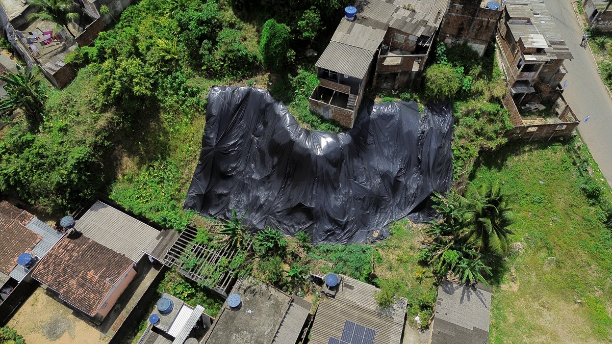 A fotografia mostra uma vista aérea de uma área urbana com casas muito próximas umas das outras. No centro da imagem, há uma grande área coberta por uma lona preta. Algumas casas têm tanques de água azuis nos telhados. Ao redor das casas, há muita vegetação verde, indicando que a região possui bastante vida vegetal.