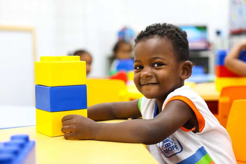 Foto em close de um menino negro, que olha para a câmera esboçando um sorriso. Ele está sentado junto a uma mesa, segurando brinquedo de blocos amarelos e azuis e usando uma farda branca com detalhes coloridos da Prefeitura do Recife.