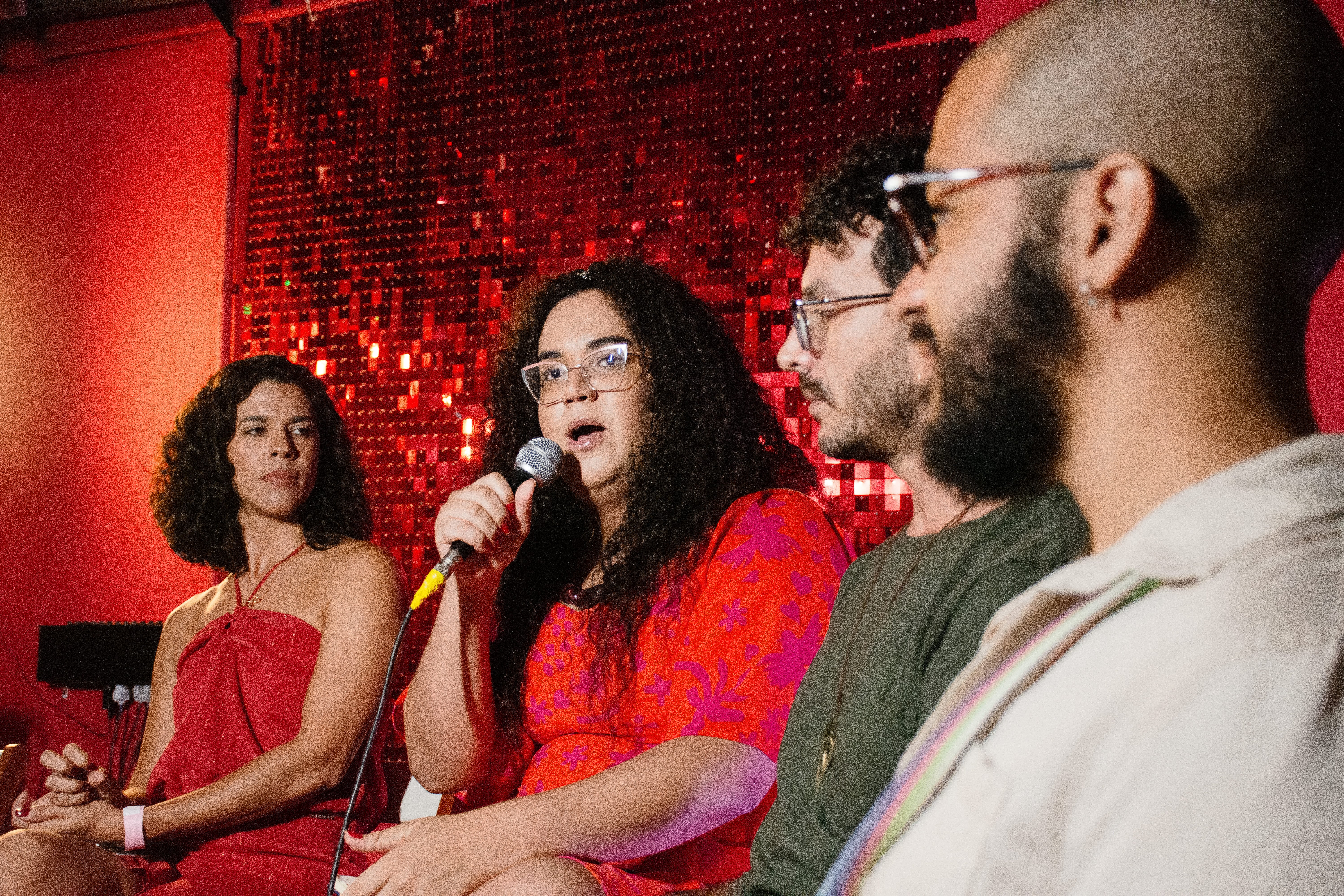 Foto de quatro pessoas sentadas, retratadas da cintura para cima em um ambiente de paredes vermelhas, com painel cintilante também na cor vermelha ao fundo. Na foto estão dois homens e duas mulheres. Os homens, ambos de barba, estão à direita da imagem, ambos usam óculos e têm barba, mas estão de perfil e levemente desfocados, apesar de ocuparem o primeiro plano da foto. Ao centro está uma mulher de cabelos encaracolados, de óculos, falando o microfone e usando um vestido vermelho com estampas de flores roxas. À esquerda, mais afastada da câmera, está outra mulher jovem, negra, de cabelos na altura dos ombros, também de vestido vermelho.