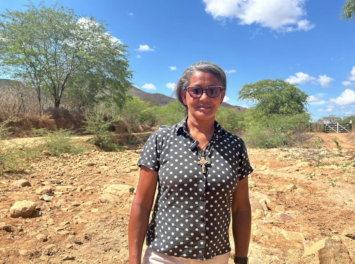 Foto de Suelina Moreira, mulher negra de cabelos grisalhos, usando óculos escuros e vestindo uma blusa preta com bolinhas brancas, com um crucifixo pendurado no pescoço. Ela foi fotografada da cintura para cima em um ambiente rural ao ar livre, em meio a um terreno pedregoso, com arbustos ao fundo e sob céu claro com poucas nuvens.