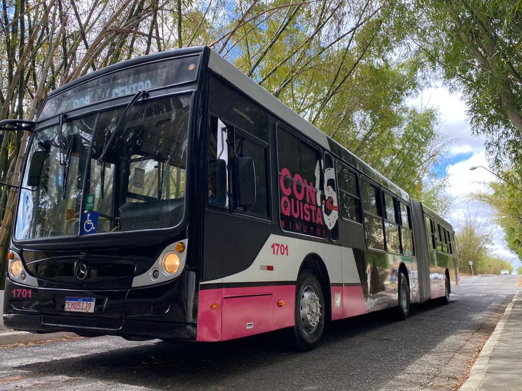 A imagem mostra um ônibus articulado grande passando em uma estrada arborizada. O ônibus é predominantemente preto com detalhes em rosa e branco. Na porta da frente, há um símbolo indicando acessibilidade para pessoas com deficiência. O nome “CONQUISTA” está escrito em letras grandes e brancas na lateral do ônibus, com “VOLVO” logo abaixo. O número do veículo “1701” está exibido acima do para-brisa e na lateral da seção traseira. A placa do ônibus também é visível. É um dia ensolarado com algumas nuvens no céu, e não há pessoas visíveis na cena.