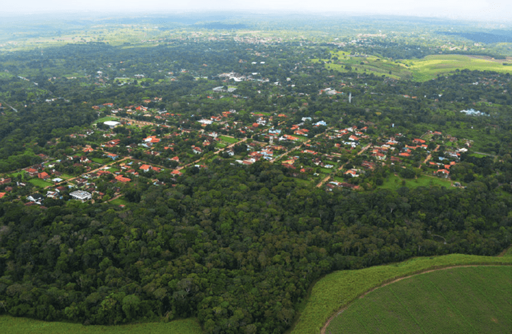 Vista aérea da APA Aldeia-Beberibe: A imagem mostra uma vista aérea de uma comunidade cercada por muita vegetação. As casas têm telhados vermelhos e estão agrupadas em algumas áreas e mais espaçadas em outras. Entre as casas, há várias árvores e espaços verdes abertos. Ao redor da comunidade, há uma floresta densa, indicando que o local está próximo ou dentro de uma grande área arborizada. Não há estradas visíveis levando à comunidade, sugerindo que pode ser um local relativamente isolado. Além da comunidade, há campos com diferentes tons de verde, possivelmente terras agrícolas, e um indício de outra comunidade distante no canto superior esquerdo.