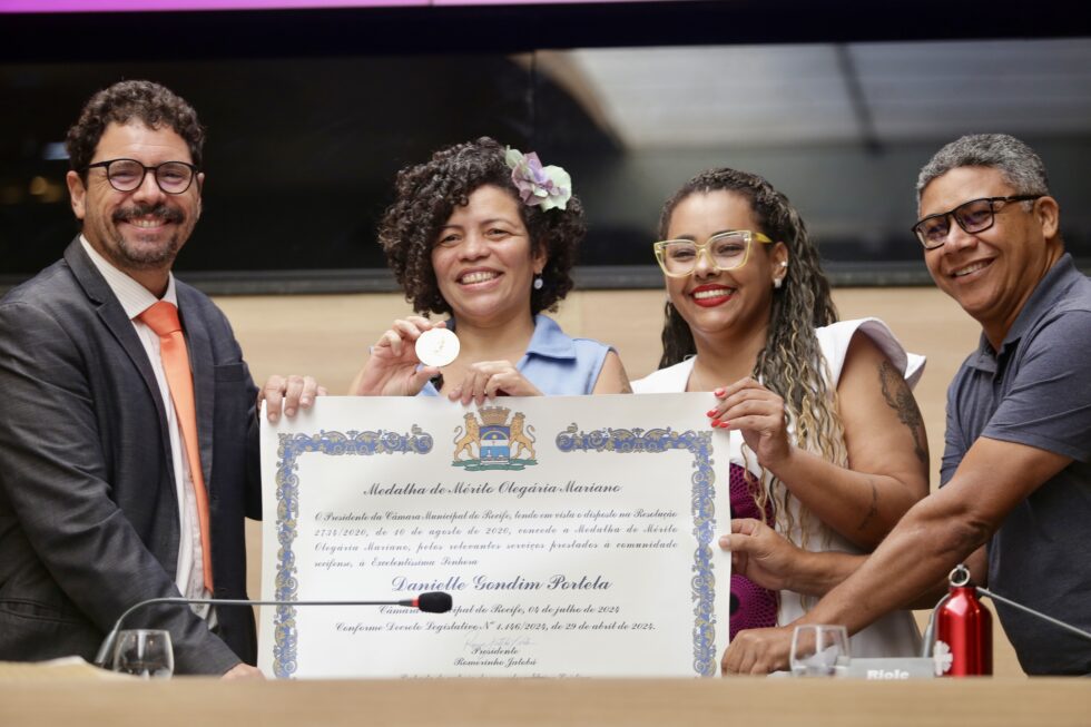 A foto mostra um grupo de quatro pessoas em pé atrás de uma mesa, segurando um grande certificado juntos. O certificado tem bordas ornamentadas e texto, mas não é totalmente visível. São dois homens, que estão nos extremos esquerdo e direito do grupo e duas mulheres negras ao centro. Eles estão vestidos de forma formal; com o homem brando da direita, usando terno, grabata laranja e óculos; as mulheres usando vestidos - uma delas com uma flor azul claro nos cabelos e o homem à direita usando uma camisa de de mangas curtas cinza chumbo. Há também um pequeno objeto metálico vermelho, parecido com um sino, sobre a mesa à frente deles. O ambiente parece ser um evento cerimonial interno, possivelmente uma cerimônia de premiação ou entrega de diplomas, indicado pelas roupas formais e pela presença do certificado.