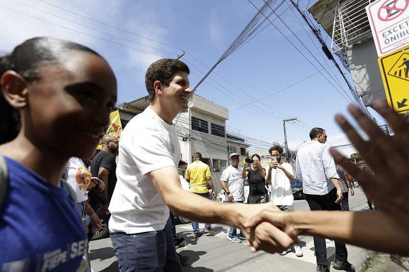 Foto de João Campos, prefeito do Recife em uma rua da periferia cumprimentando eleitores em um dia de céu azul. Ele é um homem jovem, banco, de cabelos claros, usando camisa branca de mangas curtas e calça jeans. Está cercado por pessoas, inclusive algumas que estão mais distantes que estão tirando fotos dele com celulares.