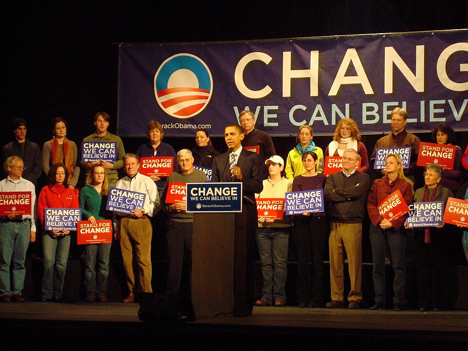 Foto de campanha de Barack Obama em pé, diante de um púlpito com microfone onde se vê um cartaz com a palavra change em primeiro plano. Por trás de Obama, estão várias pessoas de idades, gêneros e etnias diferentes segurando cartazes de campanha eleitoral.