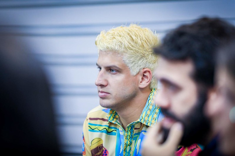 Foto de perfil de João Campos, ele é um home branco, jovem, olhos verdes, de cabelos pintados de brancos, usando uma camisa de estampado colorido. À direita da imagem, junto dele e em primeiro plano, há um outro homem, de barba e cabelos pretos, mas cujo rosto está completamente desfocado.