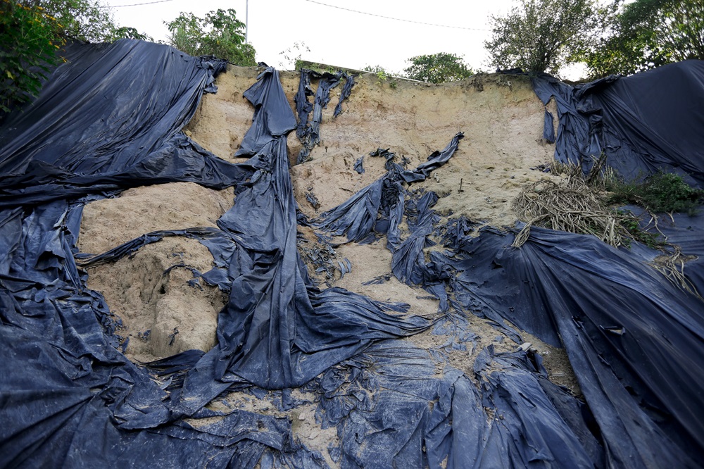 A imagem mostra uma grande encosta de terra erodida, coberta por várias peças de lona preta rasgada. A lona parece ser uma tentativa de proteger a encosta da erosão ou de conter a terra, mas está danificada e ineficaz em alguns pontos, com rasgos e buracos que deixam a terra exposta. No topo da encosta, há vegetação e uma cerca de arame ao longo da borda.