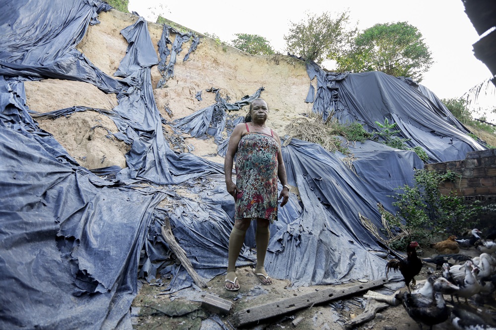 A imagem mostra uma grande encosta erodida coberta com lonas pretas. Na base da encosta, há uma mulher negra, de meia-idade, usando um vestido de estampa florida, olhando para a câmera. Ao redor dessa pessoa, há vários pedaços de madeira e destroços espalhados no chão, sugerindo uma barreira improvisada ou danos estruturais anteriores. Também há várias galinhas visíveis no primeiro plano, no canto inferior direito da foto.