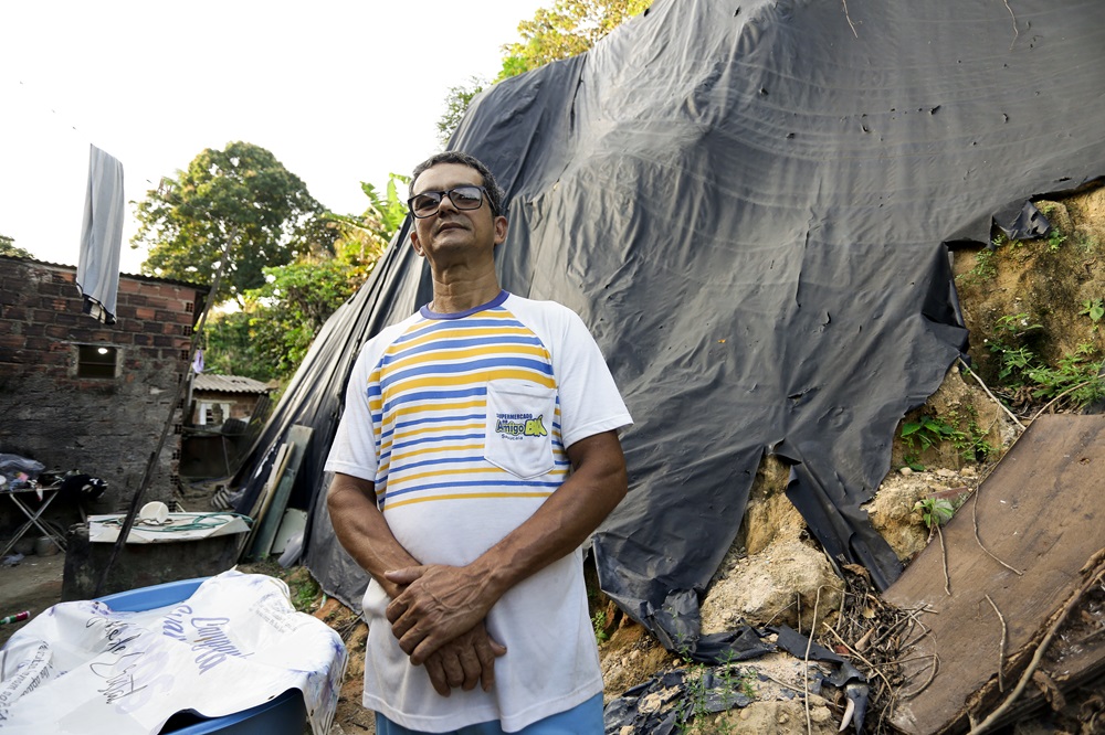 Foto de Aldemar Freire, homem de meia idade, negro e magro, usando camiseta branca com listas amarelas e azuis, posando diante de uma encosta parcialmente coberta por uma lona preta rasgada, tendo ao fundo uma construção precária, de tijolos aparentes bastante desgastados.