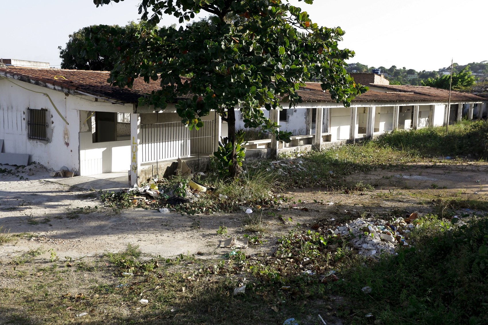 Foto de um prédio térreo, de paredes brancas, aparentando desgaste e abandono, cercado por um terreno baçdio, com montes de lixo.