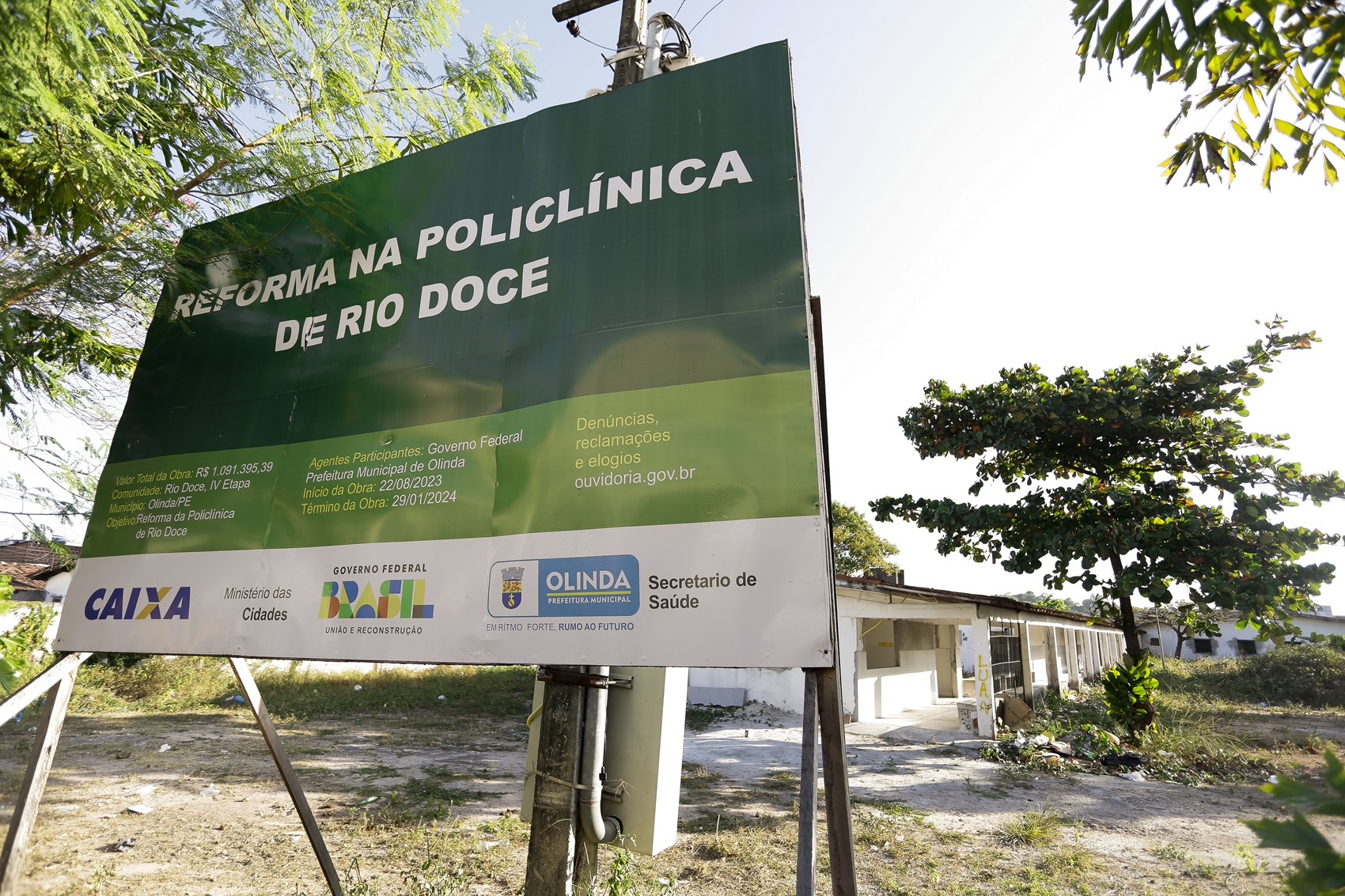 Foto da placa Reforma na policlínica de Rio Doce, diante de prédio térreo de paredes brancas e aparentando desgaste. Em torno do prédio há um terreno baldio e de relevo acidentado. A foto foi tirada de dia, com céu azul pálido.