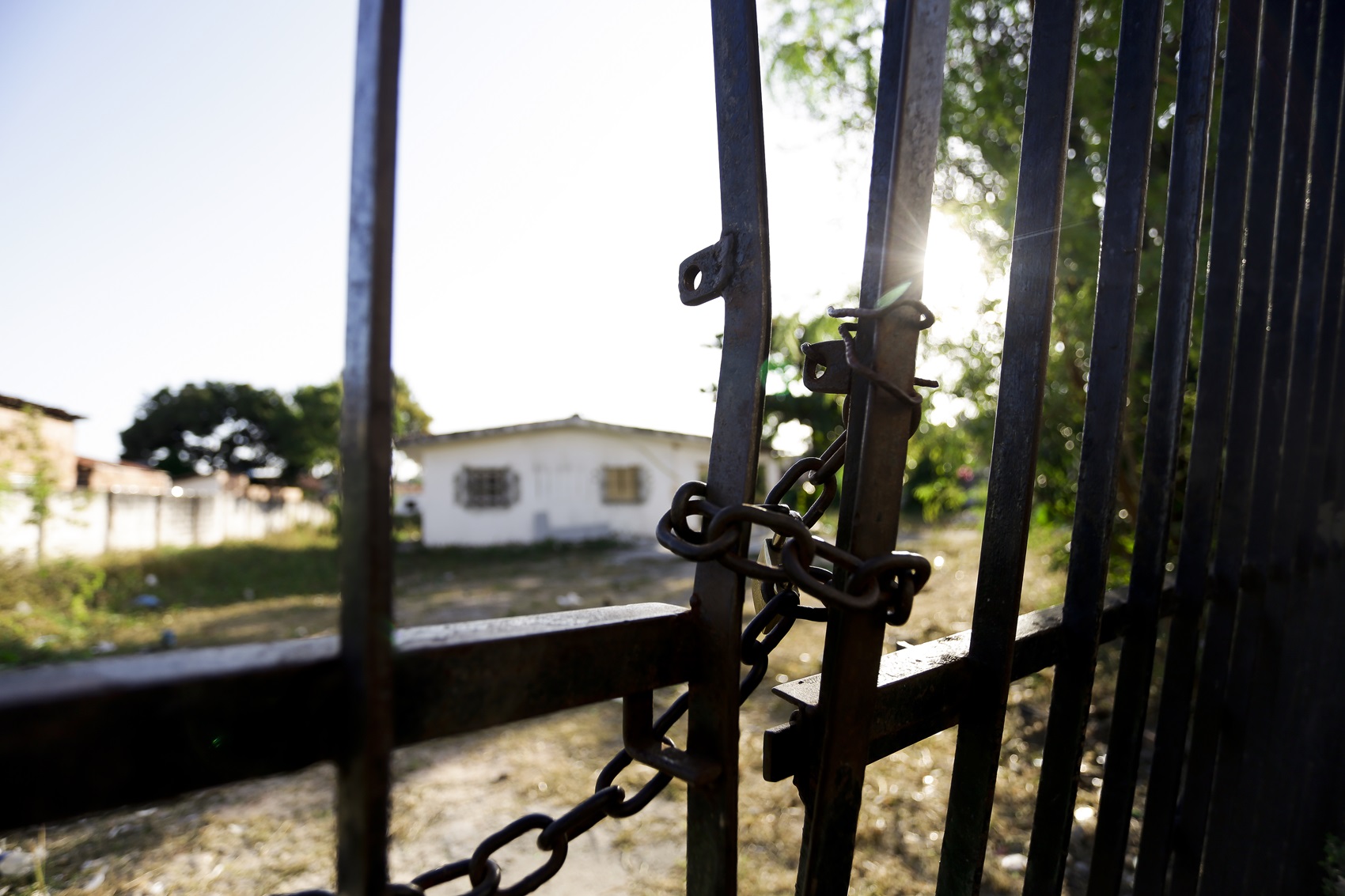 Foto de um portão de metal retorcido, com correntes, tendo ao fundo uma casa branca desgastada. A foto foi feita de dia, com sol forte e céu azul pálido.