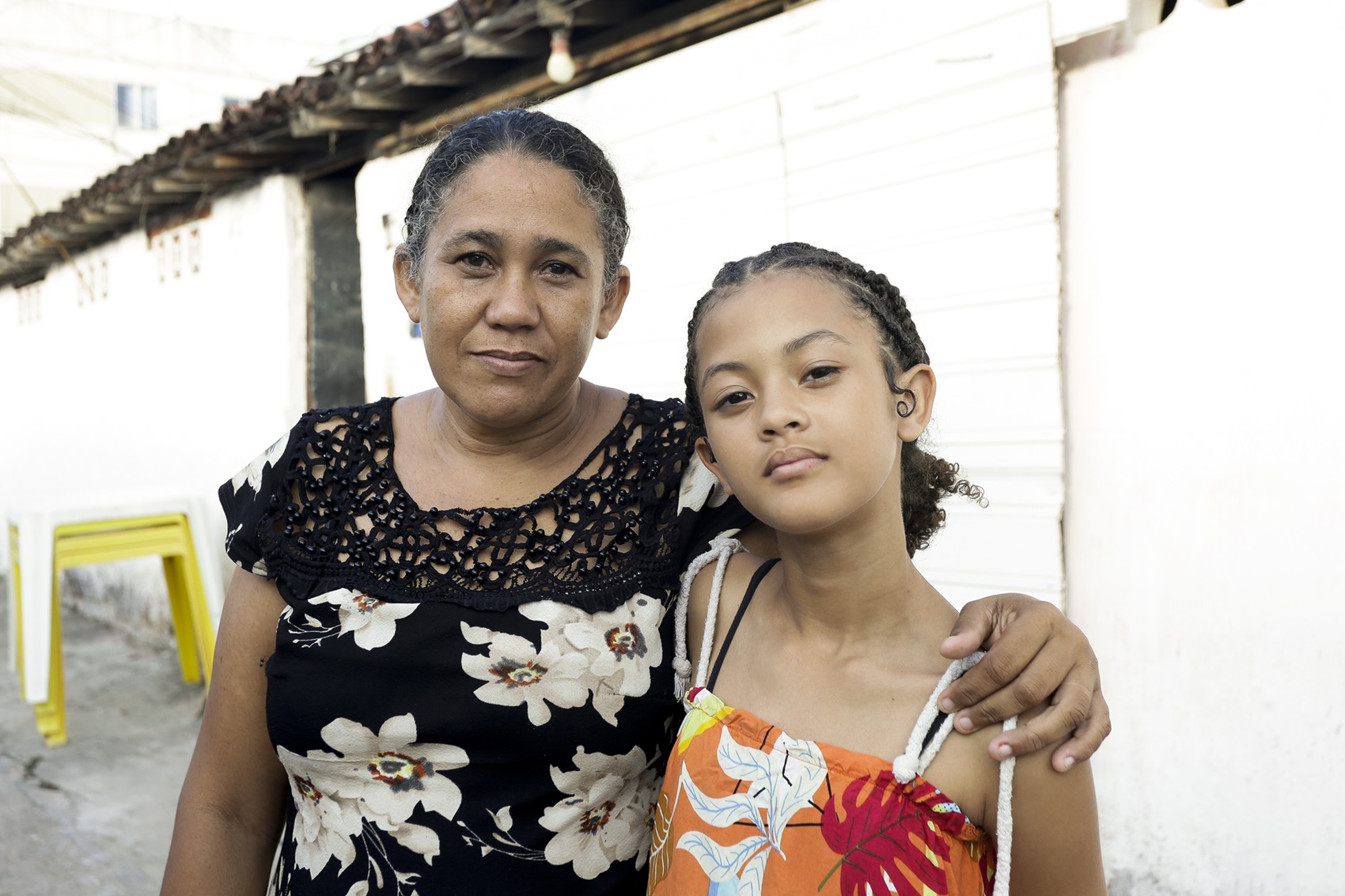 Foto de Francinete Maria Pimentel e sua filha Débora Vitória. Ela é uma mulher negra, de cabelos crespos grisalhos presos atrás da cabeça, usando um vestido preto estampado com flores brancas. A menina é negra, usando vestido estampado laranja de alças.