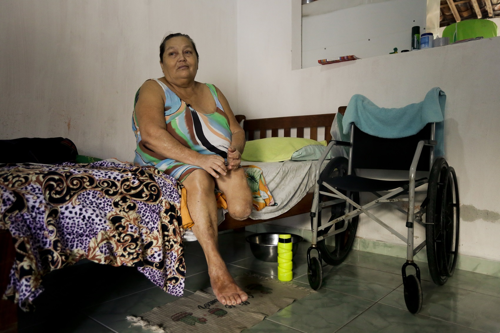 Foto de Rosilene Maria Dias, mulher parda, idosa, usando vestido estampado, de perna amputada, fotografada em um quarto de paredes brancas, cama de madeira e cadeira de rodas preta ao lado da cama.