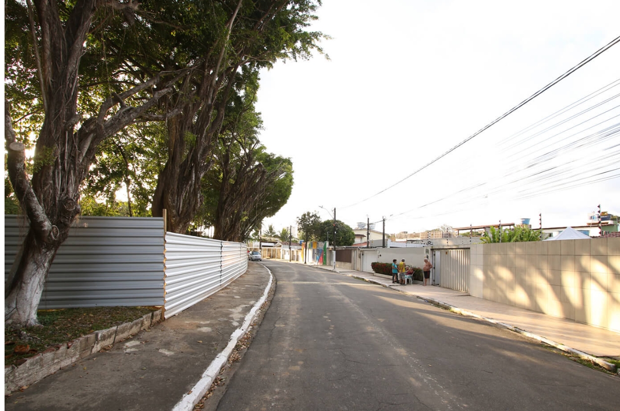 A imagem mostra uma cena urbana ao ar livre. Há uma rua que faz uma curva suave para a direita, com calçadas em ambos os lados. Do lado esquerdo da rua, há uma fileira de árvores altas com folhagem densa, proporcionando sombra sobre a calçada. Ao lado das árvores, há uma cerca alta de metal pintada com listras brancas e azuis, que parece cercar uma propriedade privada. Do lado direito da rua, há um longo muro de concreto sem janelas ou portas visíveis, sugerindo que pode ser a parede externa de outra propriedade privada. Ao longe, na curva da rua onde ela desaparece de vista, há duas pessoas: uma parece estar sentada em algo baixo, como um meio-fio ou um pequeno muro, enquanto realiza alguma atividade que não é clara à distância; a outra pessoa está em pé por perto. O céu acima está parcialmente nublado, mas principalmente claro, indicando luz de manhã ou fim de tarde. Há várias linhas de energia cruzando acima, conectadas a postes de utilidade que alinham um lado da rua.