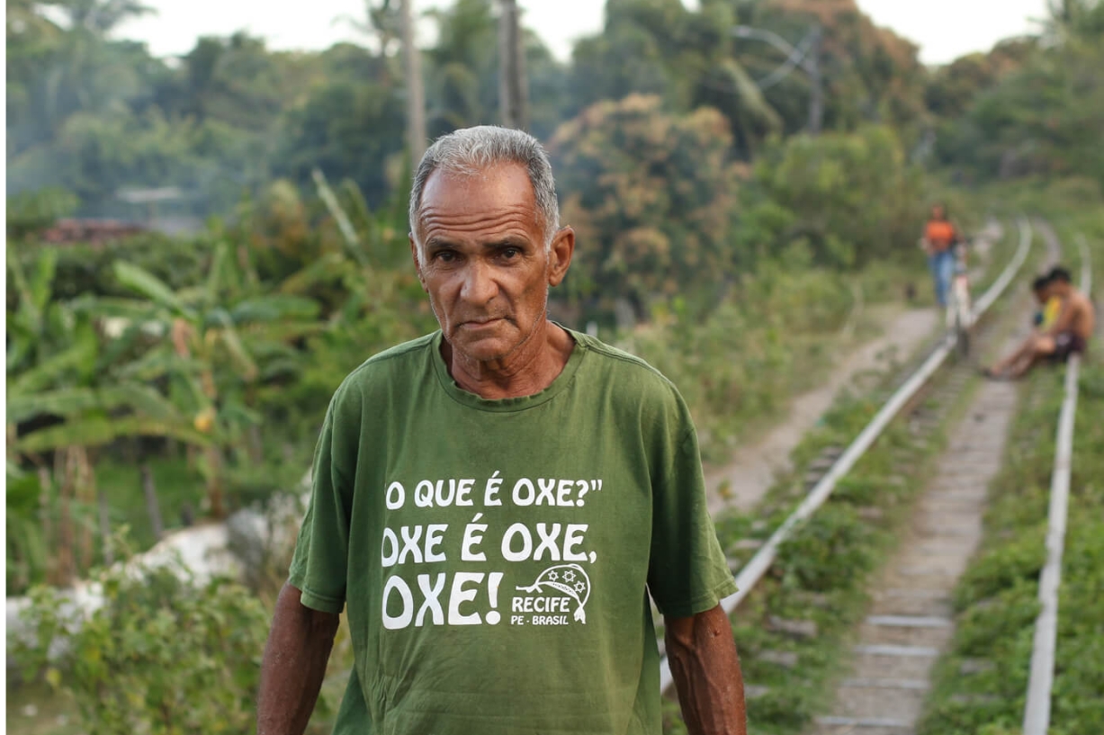 A foto mostra uma cena ao ar livre com um homem negro, de cabelos grisalhos, em primeiro plano, vestindo uma camiseta verde com a frase “O QUE É OXE? OXE É OXE, OXE!” e “RECIFE PE BRASIL,” indicando uma referência cultural a Recife, Pernambuco, Brasil. O homem olha fixamente para a câmera com semblante sério. Ao fundo, há trilhos de trem que se estendem à distância, com vegetação dos dois lados. Mais ao longe, há duas pessoas sentadas e outra em pé, possivelmente conversando ou esperando. O ambiente sugere uma área rural ou semi-rural durante o dia.