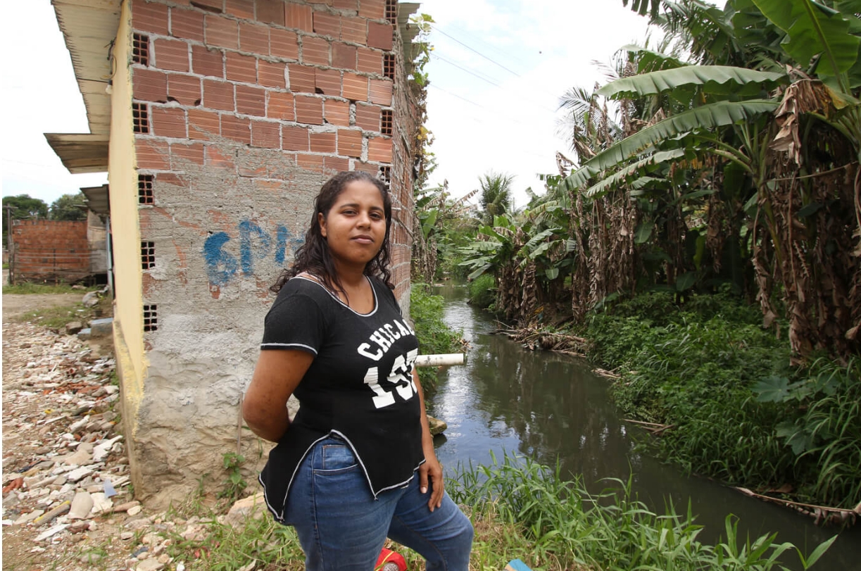 A imagem mostra uma cena ao ar livre com uma pessoa em primeiro plano. Ela é uma mulher negra, jovem, com cabelos compridos pretos, usando uma camiseta preta com detalhes brancos e calça jeans. Atrás dessa pessoa, há um canal ou riacho com água esverdeada, possivelmente indicando poluição ou estagnação. De um lado do canal, há um caminho de terra que leva a um prédio em construção, com tijolos vermelhos expostos e estrutura incompleta. Do outro lado do canal, há uma vegetação densa, incluindo bananeiras com folhas grandes.