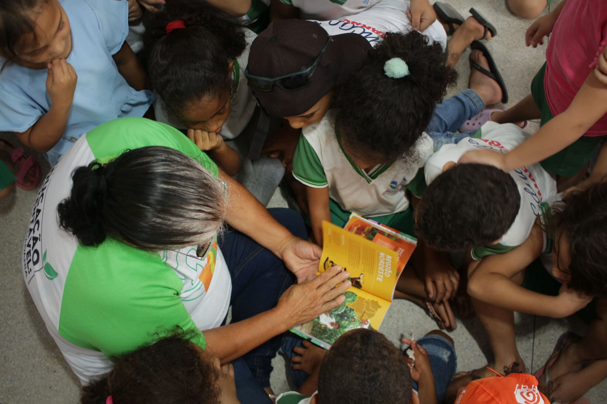 A foto mostra um grupo de crianças sentadas no chão, reunidas em volta de um adulto que está lendo um livro. O adulto parece estar apontando para algo no livro, provavelmente chamando a atenção das crianças para isso. As crianças estão usando uniformes, sugerindo que este pode ser um ambiente educacional ou uma atividade escolar. O foco da imagem está na interação entre o adulto e as crianças através da leitura.