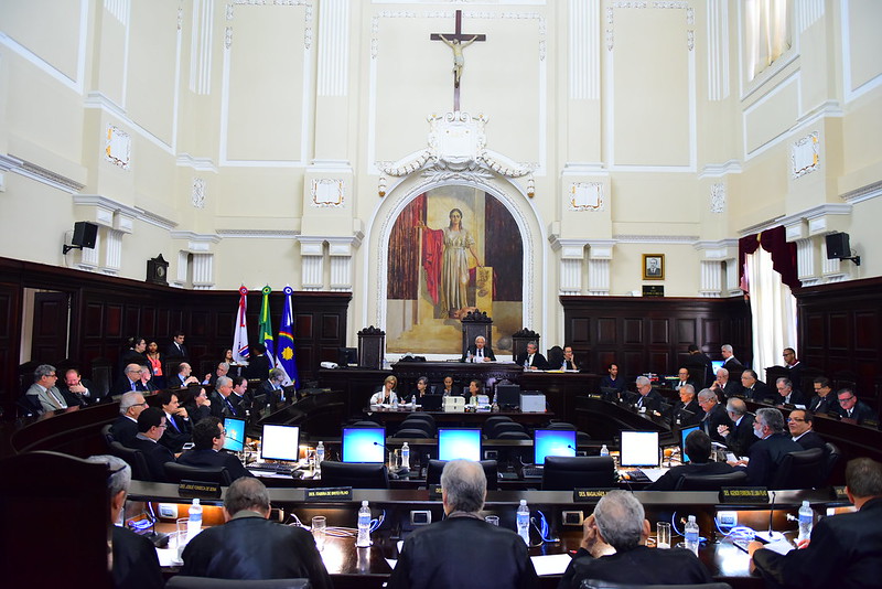 A imagem mostra o interior de um tribunal formal e grandioso. No centro, há uma grande figura de Jesus Cristo na cruz, posicionada acima de um arco. Abaixo dessa figura religiosa, há uma pessoa sentada em um banco alto, de costas para o ponto de vista, provavelmente um juiz devido ao ambiente. Essa figura central é ladeada por duas grandes pinturas, uma de cada lado do arco. A sala está cheia de várias fileiras de indivíduos vestidos com togas escuras, provavelmente advogados ou juízes, sentados em longas mesas voltadas para o banco alto e uns para os outros, de forma simétrica. As paredes são adornadas com molduras decorativas e iluminadas por lustres pendurados no teto. Há bandeiras exibidas em ambos os lados da sala, perto da frente.