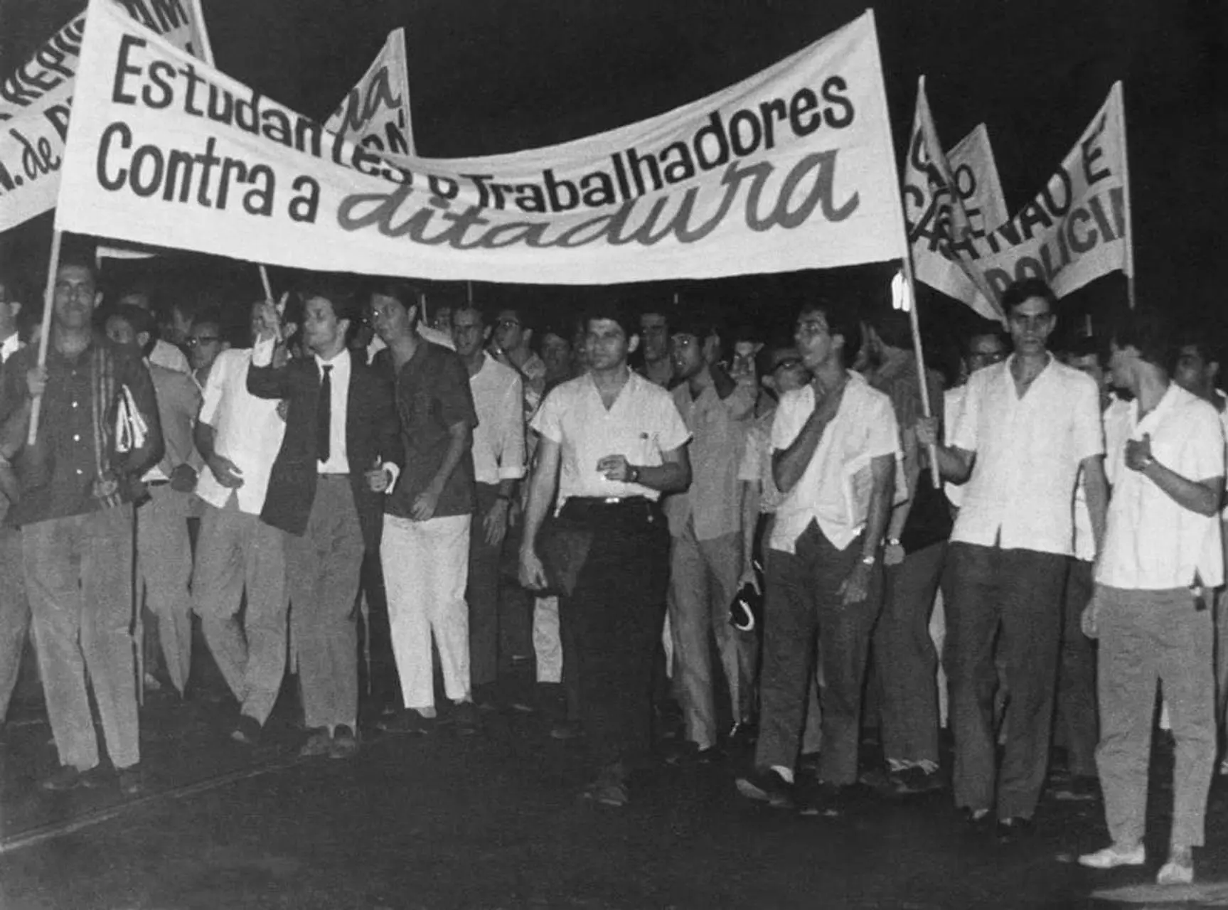 A foto em preto e branco mostra um grupo de pessoas participando de um protesto ou manifestação. Elas estão segurando faixas com a frase “Estudantes e Trabalhadores Contra a Ditadura.” A imagem captura um momento de ação coletiva e resistência contra um regime autoritário, refletindo movimentos sociais e políticos históricos.