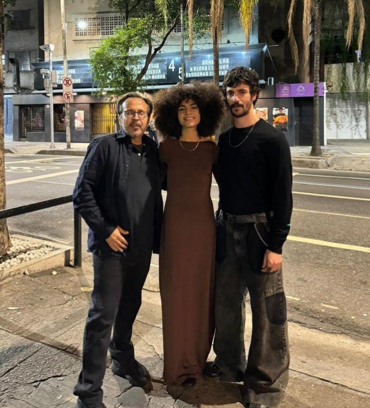 Foto noturna de Rafael Conde, Samantha Jones e Caio Horowicz na rua, em frente a um cinema. Rafael é um homem de meia idade, cabelos castanhos claros, de óculos, bigode e barba ralos, usando jaqueta preta, camisa preta e calça cinza escura; Samantha é uma mulher jovemm negra, de fartos cabelos cacheados, usando vestindo longo na cor bordô; Caio é um homme jovem, magro, de cabelos curtos pretos e barba e bigode ralos. Ele usa uma camisa preta de mangas compridas e calça larga e cinza.