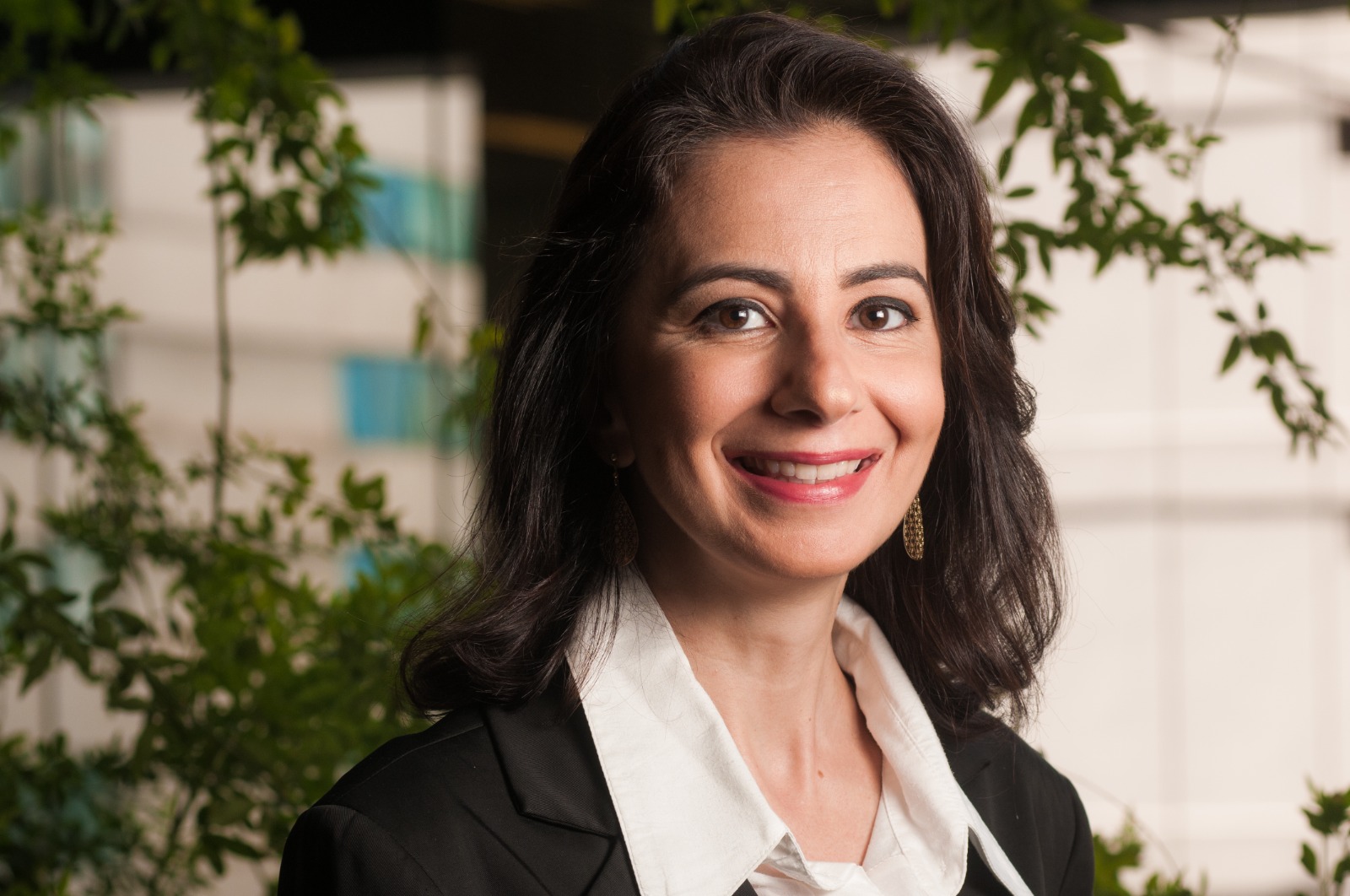 Foto em close de Beatriz Abuchahim, mulher branca, de meia idade, cabelos castanhos compridos, usando blazer preto e blusa branca. Ela está sorrindo para a câmera e foi fotograda em ambiente fechado, tendo ao fundo plantas em vasos no chão e na parede.