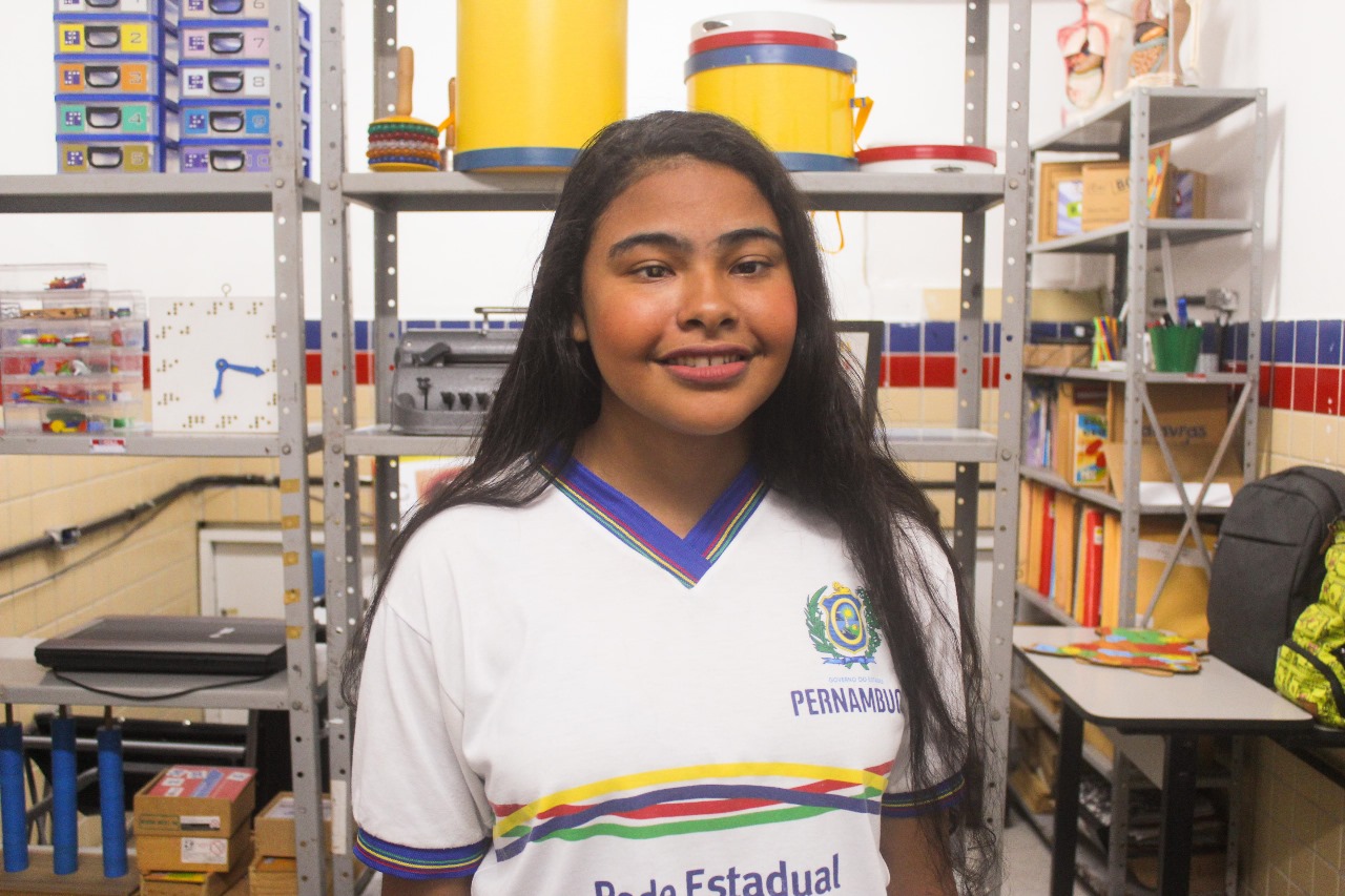 Foto de uma menina negra, de cabelos pretos, lisos e compridos. A menina na foto parece estar em uma sala de aula ou em um depósito. No fundo, há estantes de metal cheias de vários itens, como livros, recipientes de armazenamento e materiais educativos. As estantes estão bem organizadas, e há um recipiente amarelo na prateleira superior que se destaca pela cor. A menina está vestindo um uniforme branco com detalhes em azul, verde, vermelho e amarelo, que inclui um emblema de Pernambuco.