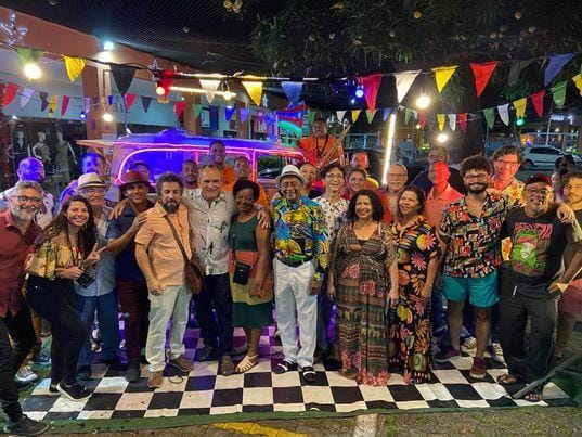 Homens e muheres com roupas multicoloridas posando para a foto à noite em um ambiente externo decorado no alto nom bandeirinhas coloridas triangulares usadas em festas juninas. O grupo está pisando em uma espécie de tapete quadriculado em preto e branco, tendo ao fundo uma estrutura colorida de luz neon.