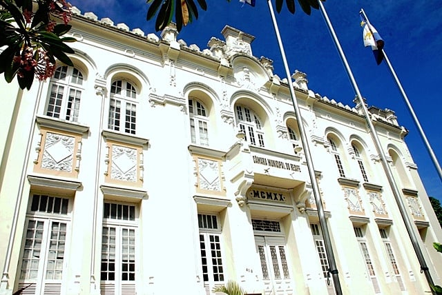 A imagem mostra um edifício histórico e grandioso com uma fachada ornamentada, apresentando paredes brancas e molduras decorativas. A arquitetura inclui janelas arqueadas com grades intrincadas e uma entrada central ladeada por duas colunas. Acima da entrada, há uma placa que diz “CÂMARA MUNICIPAL” seguida por “MCMXIX,” que indica o ano de 1919 em algarismos romanos. O edifício é adornado com bandeiras em mastros na frente dele, e há plantas verdes exuberantes parcialmente visíveis nas laterais. O céu está claro e azul, sugerindo um dia ensolarado.